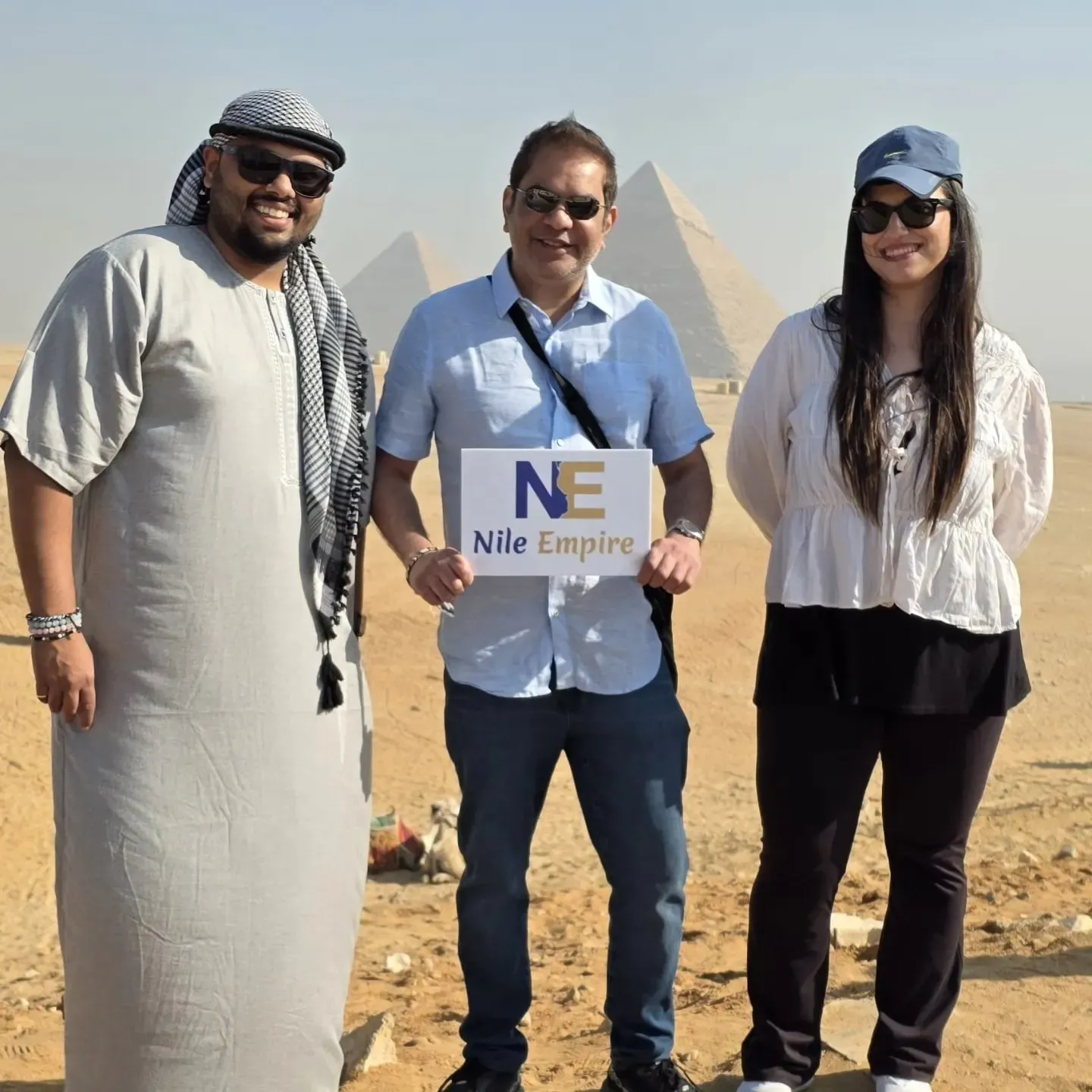Tres visitantes sonrientes con un cartel de Nile Empire frente a las pirámides de Giza