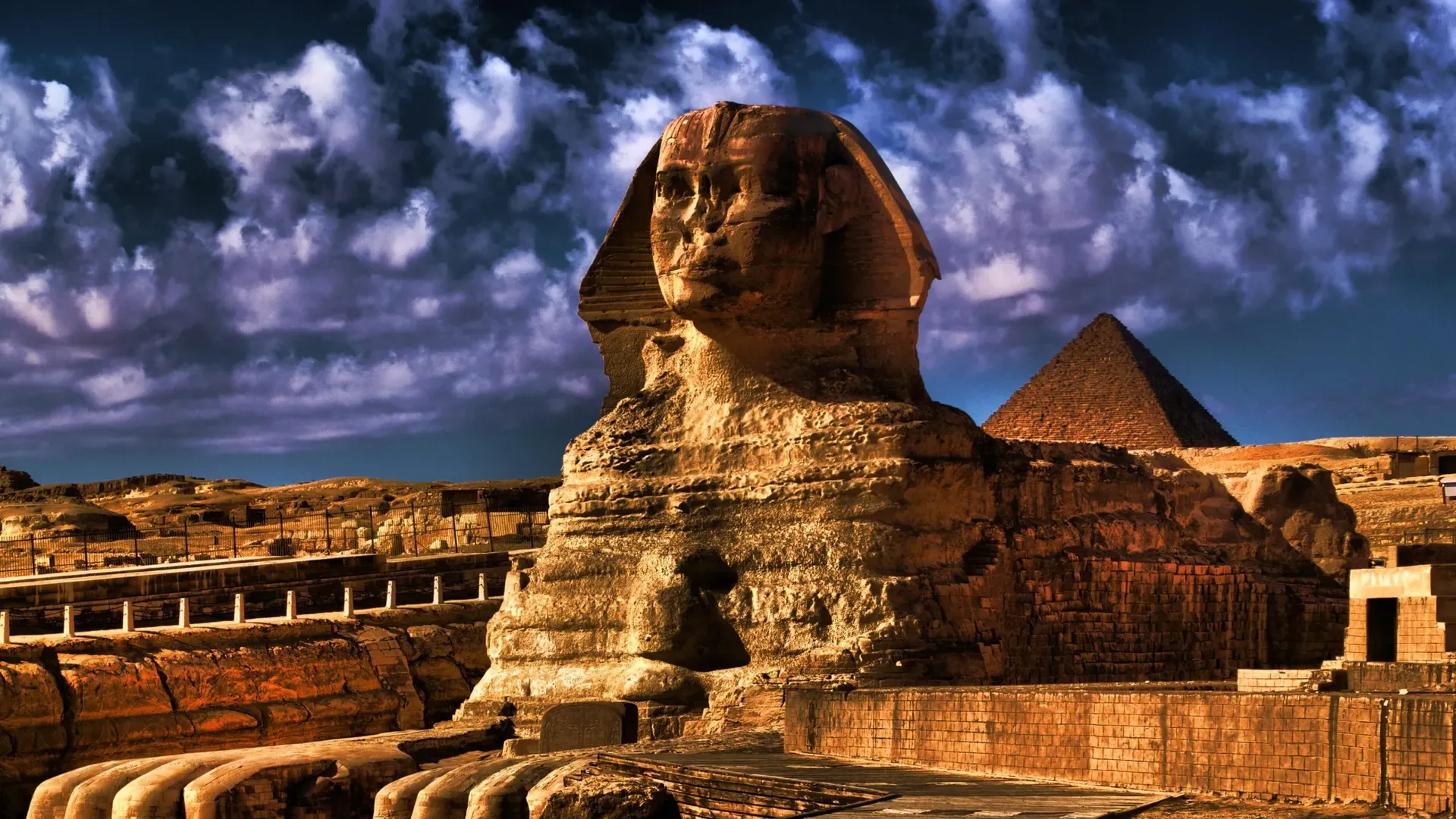 La Gran Esfinge de Giza en primer plano, con una pirámide detrás y un cielo de nubes dramáticas