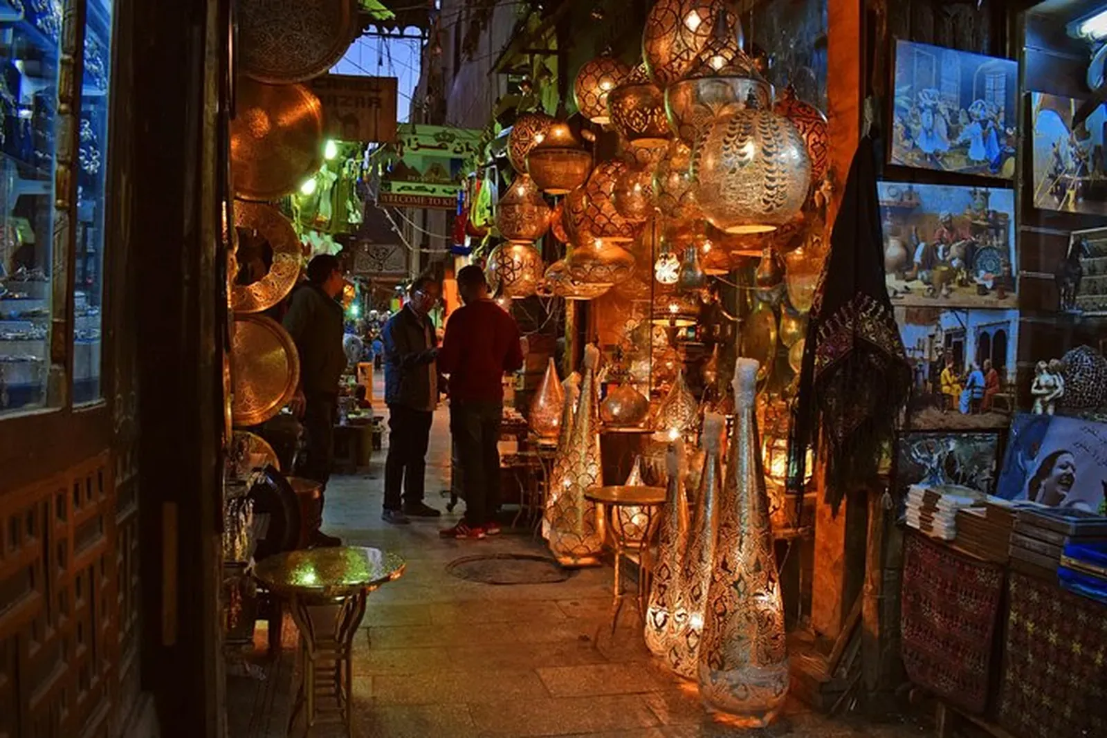 Lámparas de latón encendidas a lo largo de un estrecho callejón de bazar al anochecer, con algunos compradores